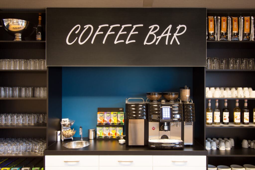 Projectkeuken koffiebar met koffiemachine en servies in zakelijke omgeving. Inclusief wandbelettering.